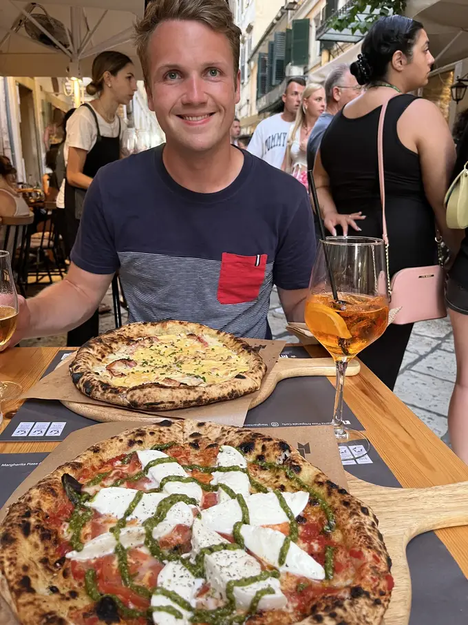 Max an einem Tisch in einer Gasse, vor ihm zwei Pizzen und ein orangefarbenes Getränk im Weinglas