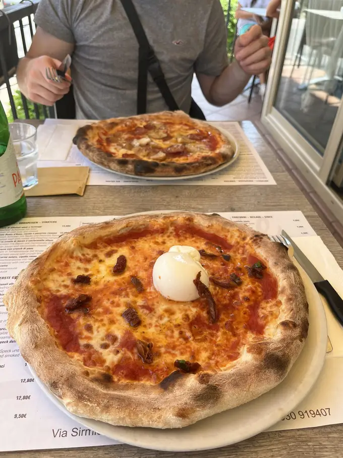 Zwei Pizzen auf einem Tisch mit Papierset, vorne eine mit Burrata und getrockneten Tomaten, dahinter Max im grauen T-Shirt mit Besteck in der Hand