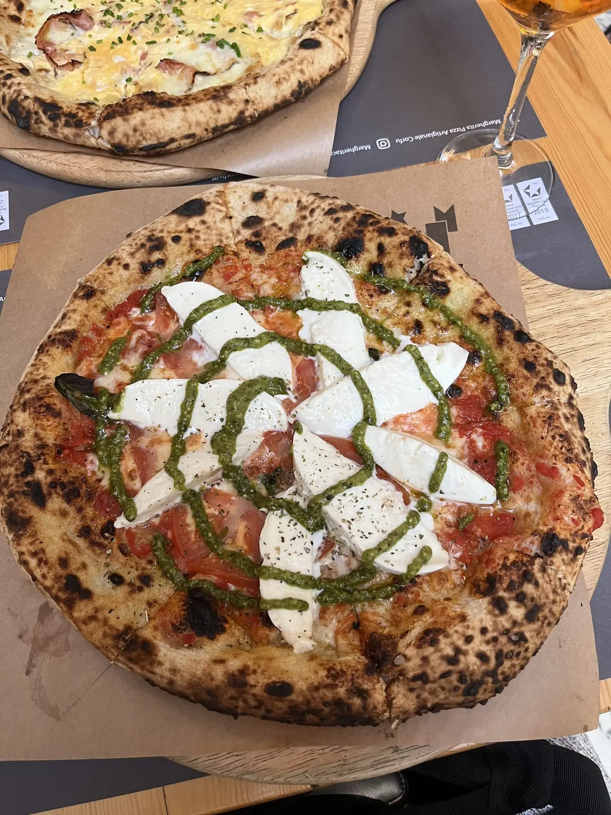Pizza mit Mozzarella und Pesto auf einem Holzbrett, dahinter eine zweite Pizza und ein Weinglas