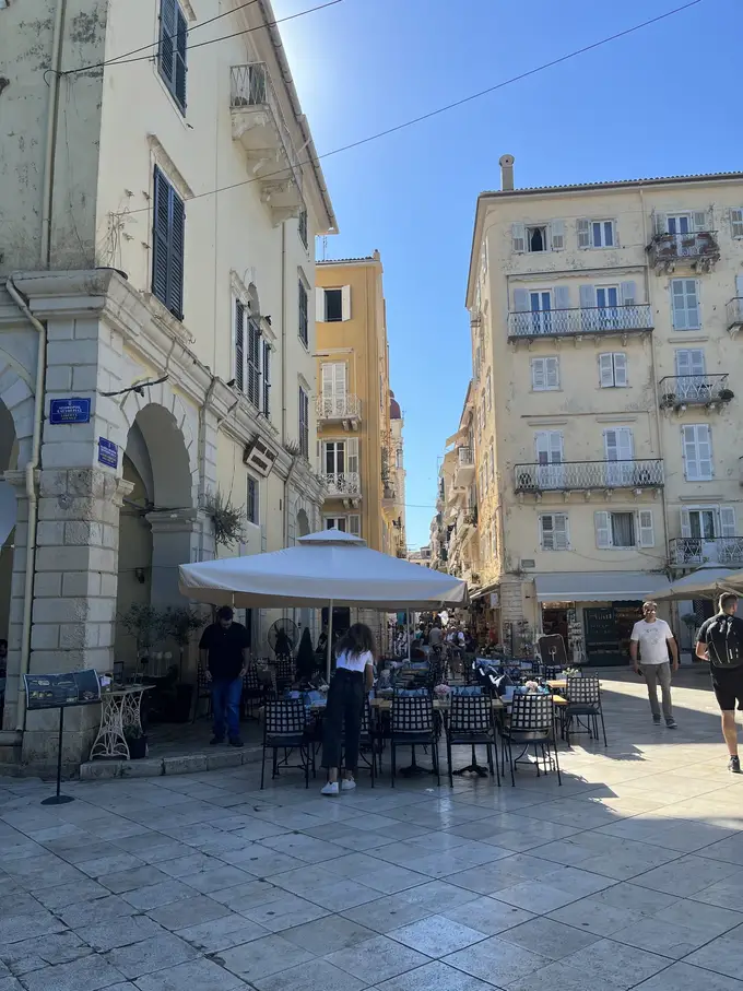 Kleiner Platz mit Café-Tischen unter einem Sonnenschirm, dahinter hohe Häuser mit Fensterläden