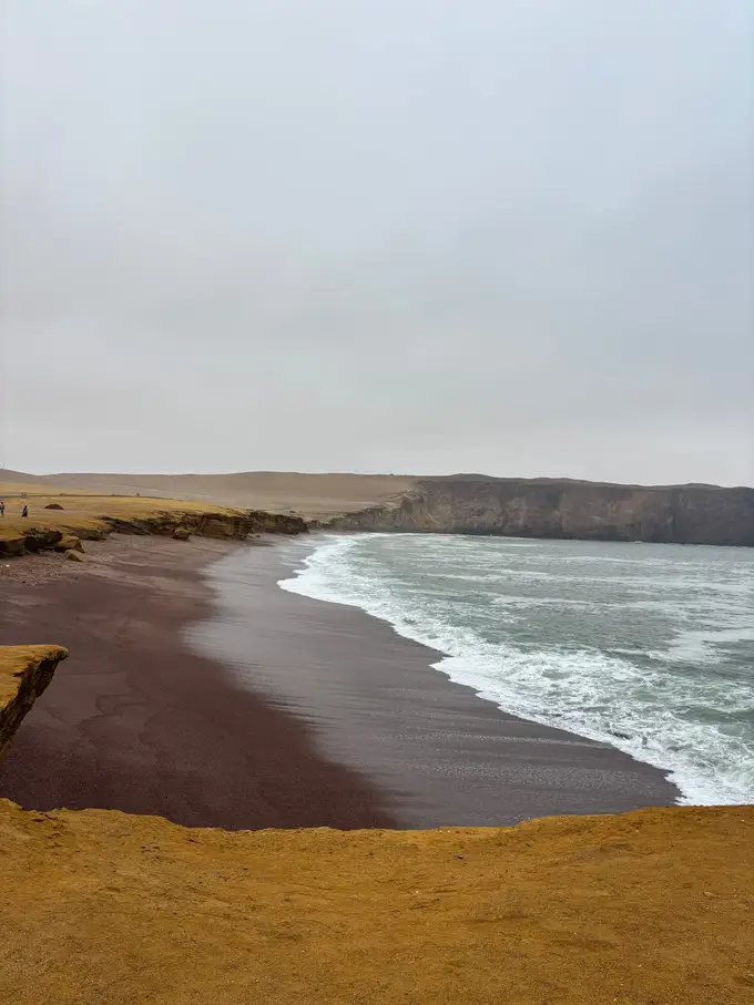 Ein rotbrauner Sandstrand unter einer ockerfarbenen Abbruchkante, die Wellen laufen weiß schäumend auf