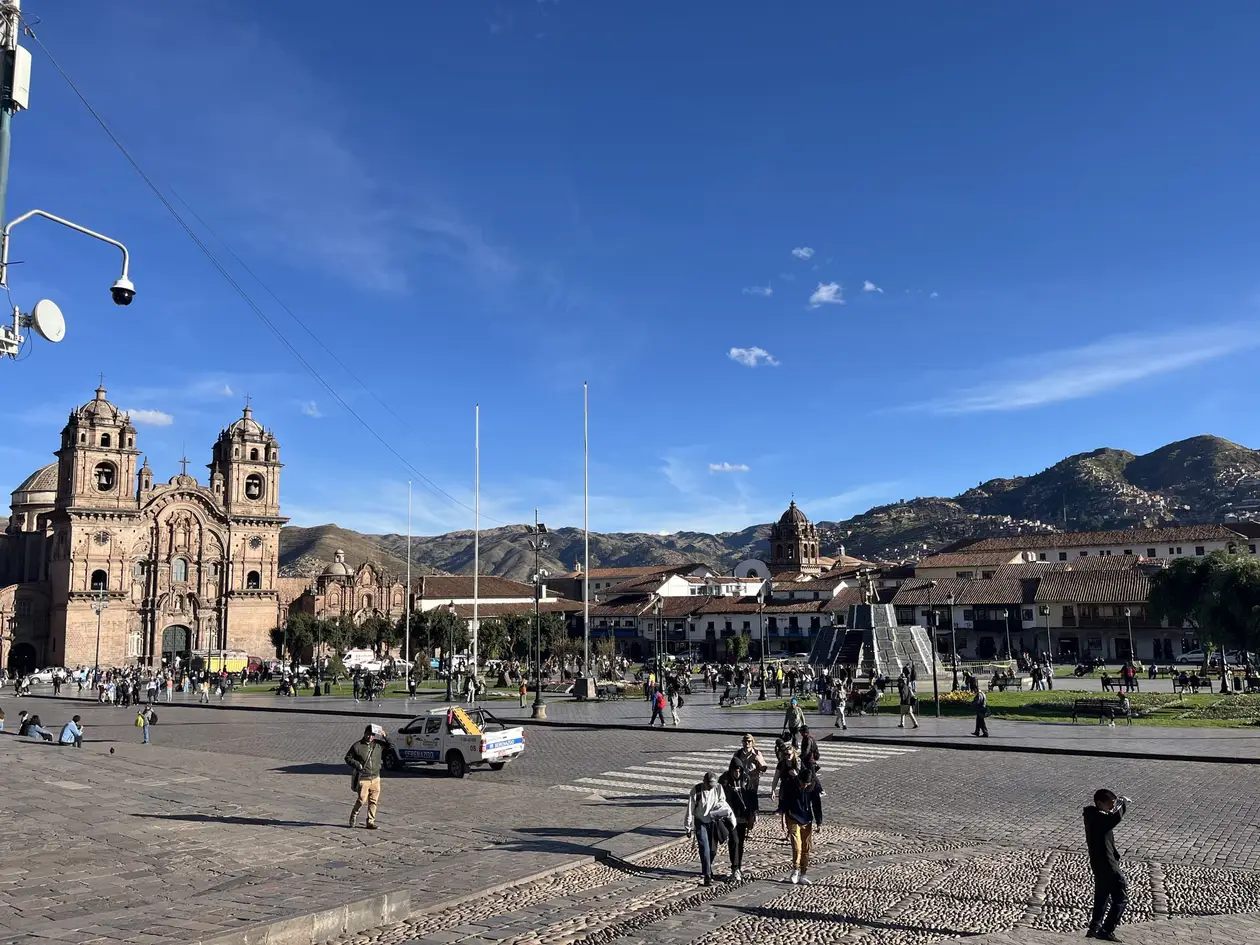 Die Plaza de Armas von Cusco unter blauem Himmel, links die Kirche mit zwei Türmen, dahinter die Hügel der Stadt