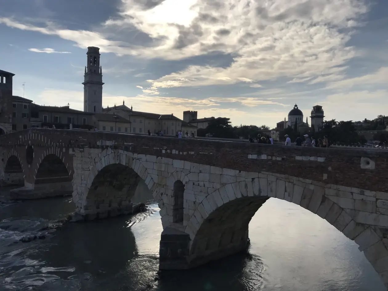 Die steinerne Bogenbrücke Ponte Pietra über die Etsch im Abendlicht, dahinter ein Glockenturm und Kuppeln
