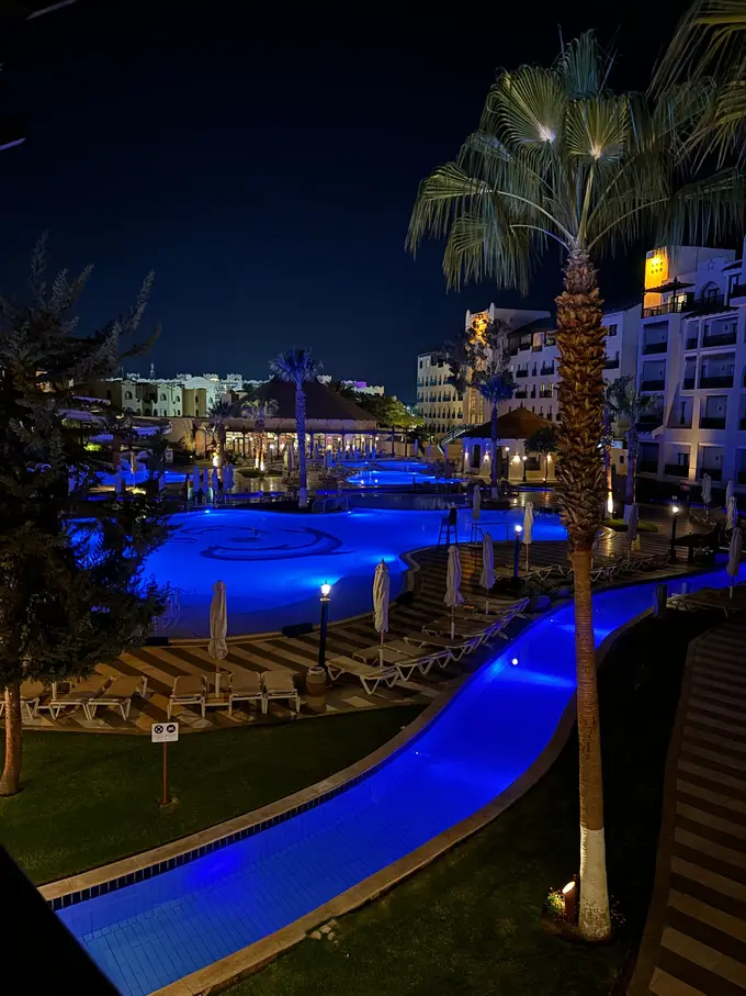 Die Poolanlage bei Nacht, das Wasser blau beleuchtet, daneben Palmen, leere Liegen und angestrahlte Hotelgebäude