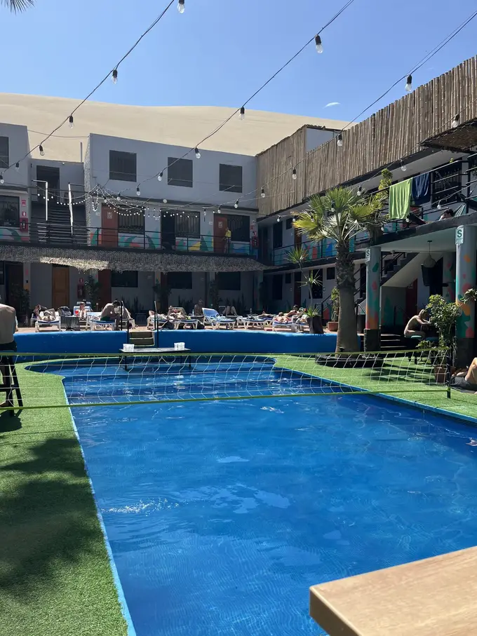 Blauer Pool mit Kunstrasen im Innenhof des Hostels, ringsum zweistöckige Zimmertrakte und Liegen, dahinter eine Düne