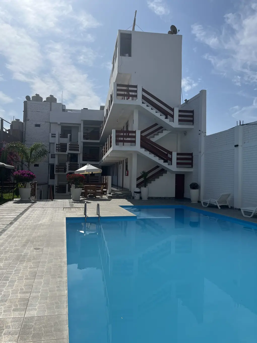 Hellblauer Pool in einem gefliesten Innenhof, dahinter ein weißes mehrstöckiges Haus mit roten Außentreppen