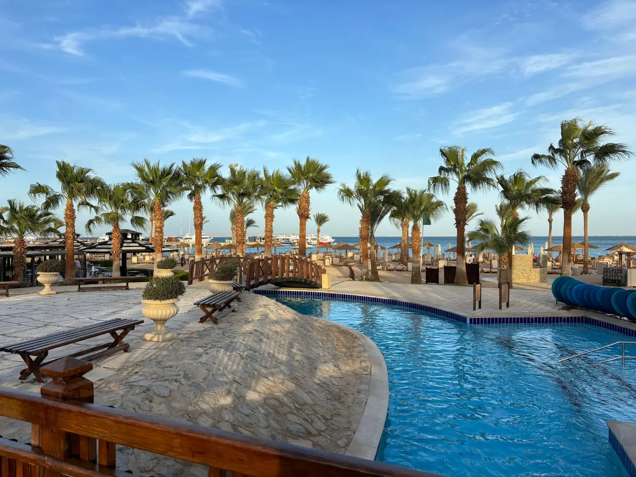 Ein geschwungenes Poolbecken mit Holzbrücke, dahinter eine Reihe Palmen, Sonnenschirme aus Stroh und das Meer mit Ausflugsbooten