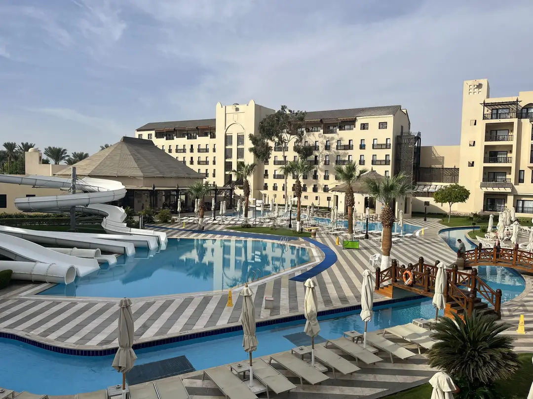 Blick über die Poolanlage mit weißen Wasserrutschen, gestreiften Terrassen, Holzbrücke und hellen Hotelgebäuden, die Liegen sind noch leer