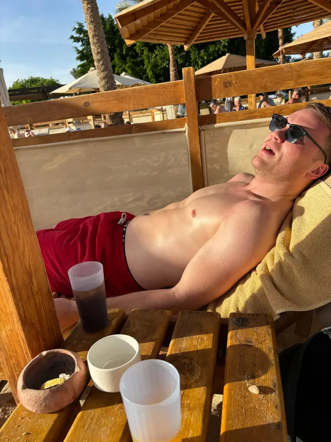 Max liegt mit Sonnenbrille und offenem Mund auf einer Liege unter einem Strohschirm, davor ein Tisch mit Getränken und einem Aschenbecher