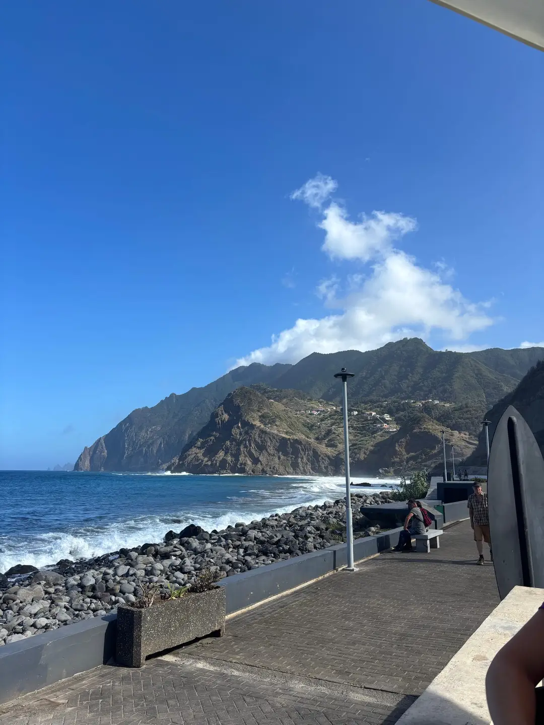 Gepflasterte Uferpromenade neben dunklen Lavafelsen, dahinter die Küste und ein Ort am Hang