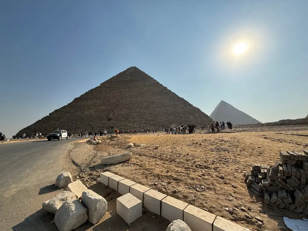 Eine große Pyramide an einer Straße mit Polizeiwagen, davor Besuchergruppen, rechts dahinter eine zweite Pyramide, im Vordergrund Pflastersteine und Randsteine