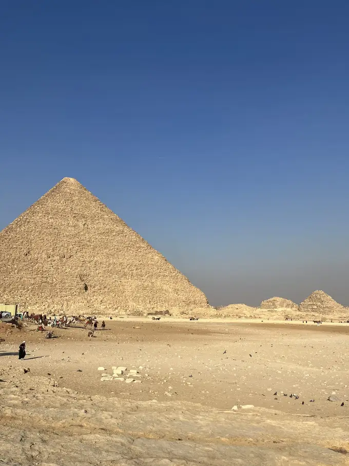 Eine große Pyramide unter tiefblauem Himmel, rechts daneben kleinere, verfallene Pyramiden, davor eine Sandfläche mit Kamelen, Reitern und Tauben
