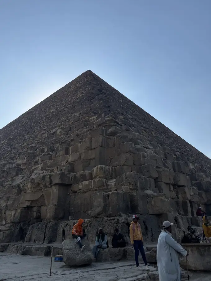 Die Ecke einer Pyramide von unten gegen den Himmel, am Fuß sitzen und stehen Männer auf den untersten Steinblöcken