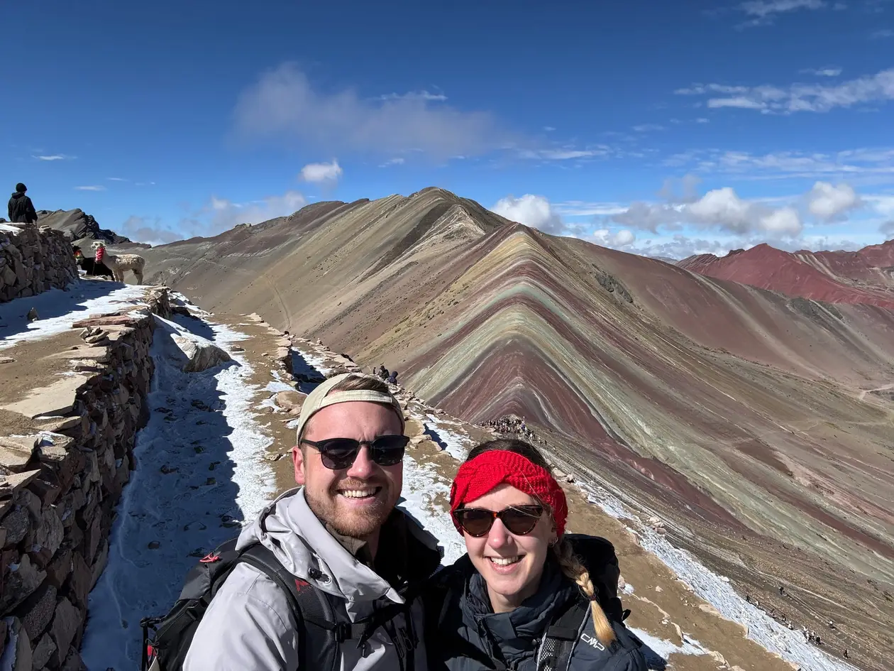 Selfie von Max und Rebecca auf dem Grat, dahinter der Rainbow Mountain mit seinen farbigen Gesteinsstreifen und Schnee am Weg