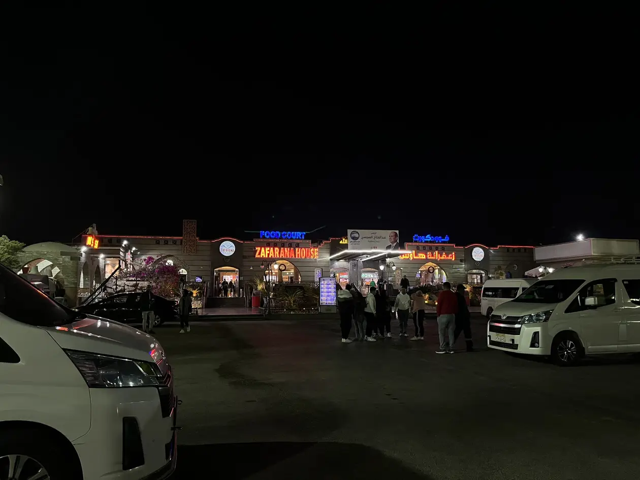 Eine beleuchtete Raststätte bei Nacht mit Leuchtschriften, davor ein Parkplatz mit weißen Kleinbussen und einer Gruppe Reisender