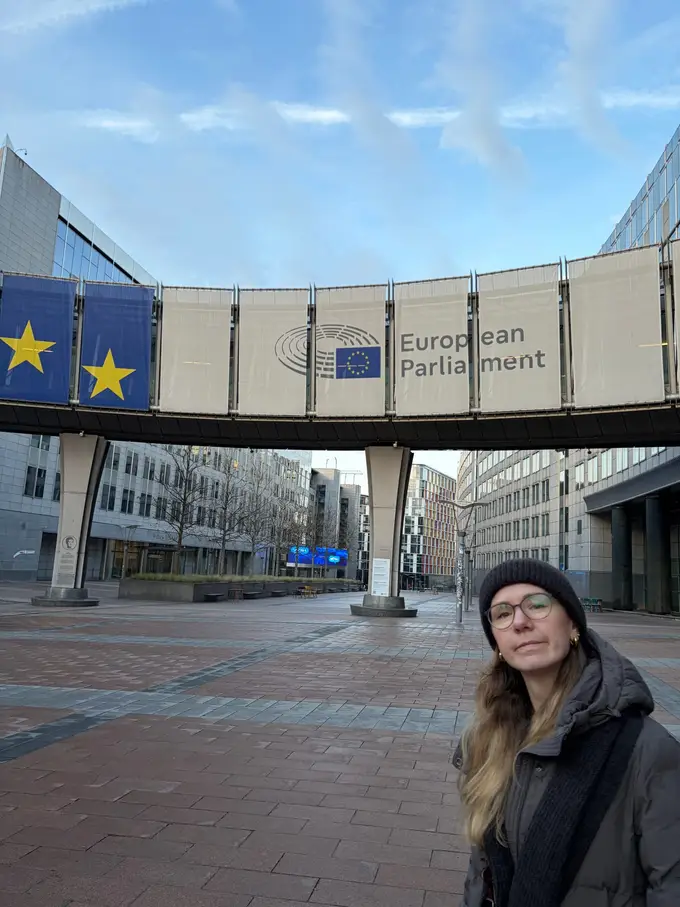 Rebecca vorne rechts im Bild, hinter ihr ein Übergang zwischen zwei Gebäuden mit der Aufschrift European Parliament und Europaflaggen