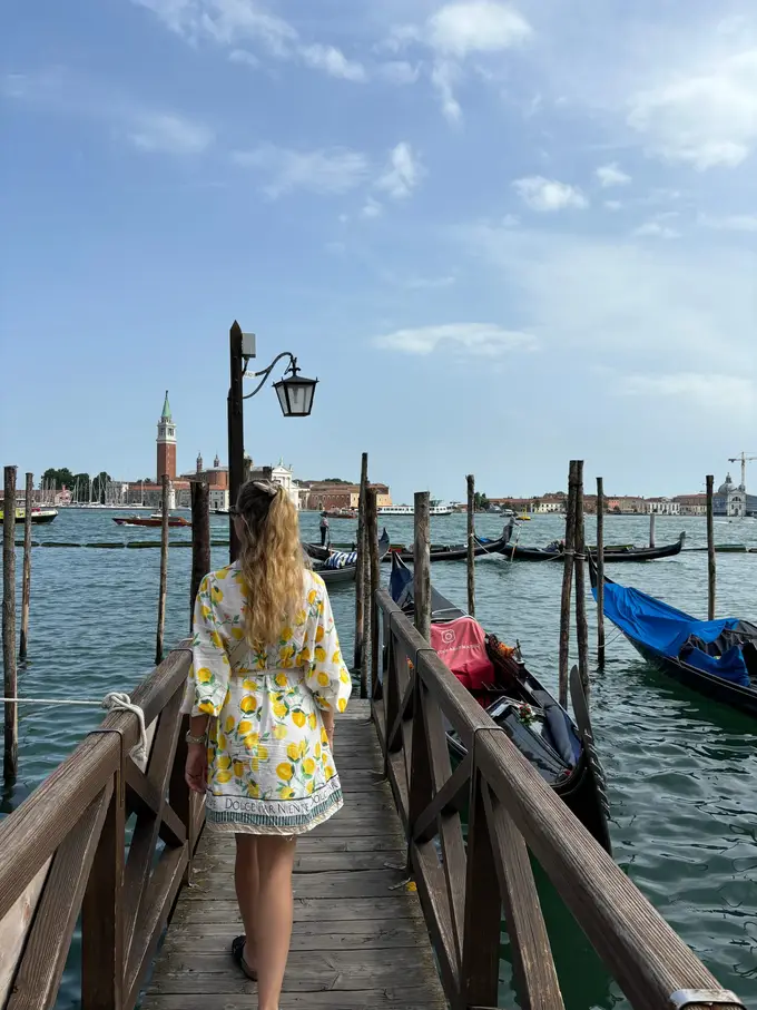 Rebecca im gelb-weißen Zitronenkleid geht von hinten gesehen über einen Holzsteg zwischen Gondeln, gegenüber die Insel San Giorgio Maggiore mit Kirchturm