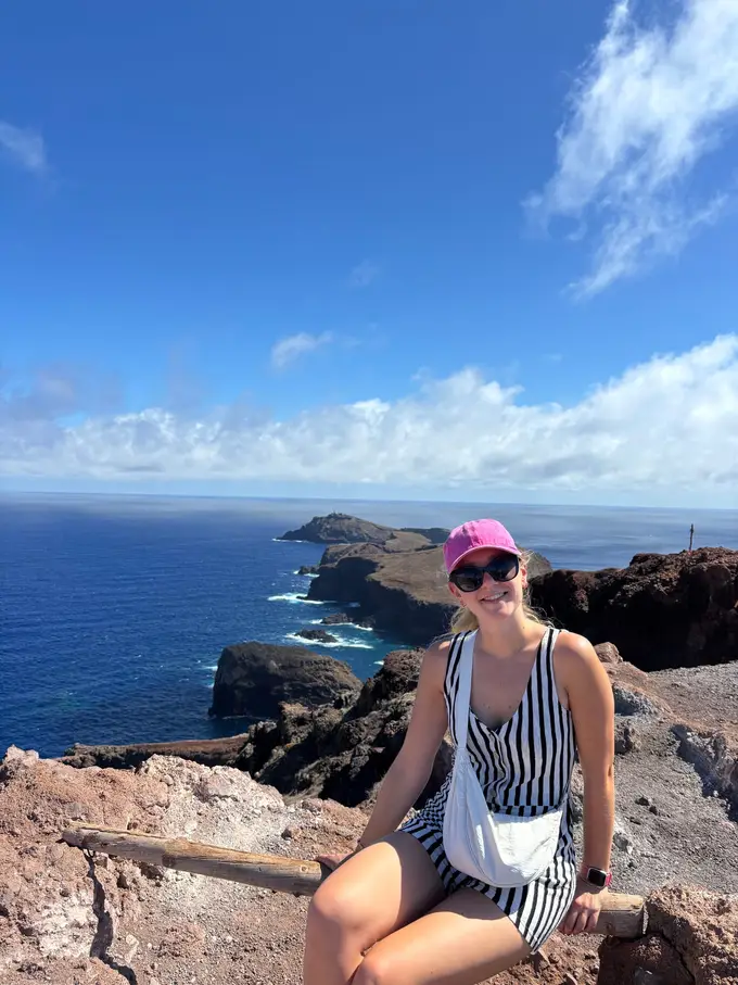 Rebecca sitzt mit Käppi und Sonnenbrille auf Felsen, dahinter in praller Sonne die zerklüftete Küste und andere Wanderer