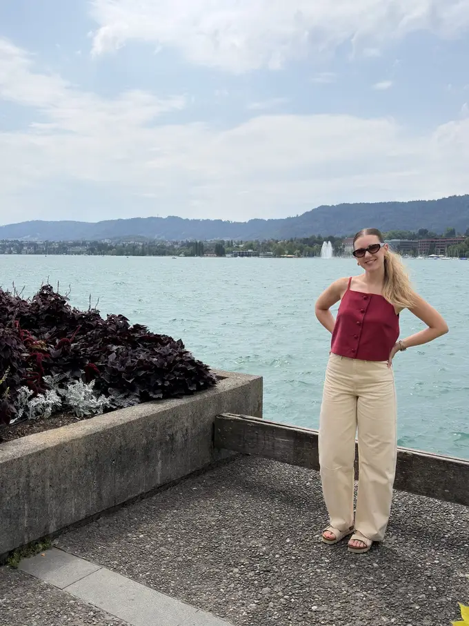 Rebecca steht mit den Händen in den Hüften am Ufer des Zürichsees, hinter ihr türkises Wasser, eine Fontäne und bewaldete Hügel