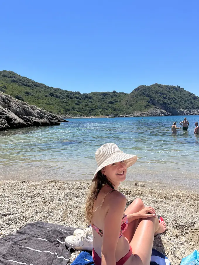 Rebecca mit Sonnenhut sitzt auf einem Handtuch am Kiesstrand, dahinter klares Wasser und grüne Hügel