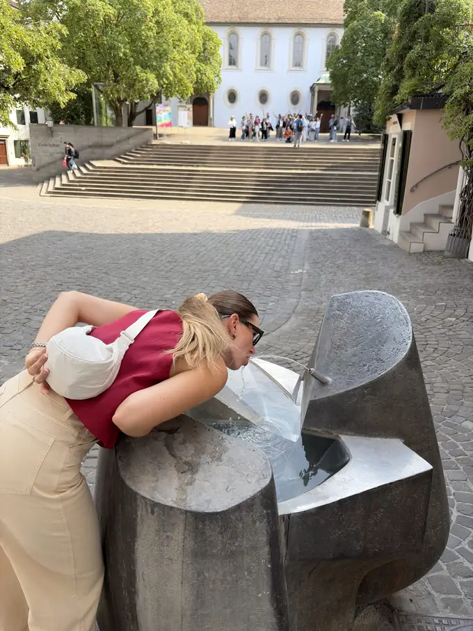 Rebecca beugt sich über einen modernen Steinbrunnen und trinkt aus dem Wasserstrahl, dahinter eine breite Treppe und eine Kirche