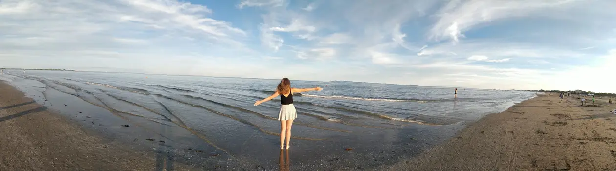 Panorama eines flachen Sandstrands, Rebecca steht mit ausgebreiteten Armen im seichten Wasser