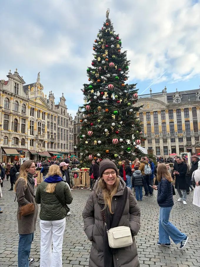 Rebecca in Daunenmantel und Mütze lacht auf dem Grand-Place, hinter ihr ein großer geschmückter Weihnachtsbaum und die Zunfthäuser
