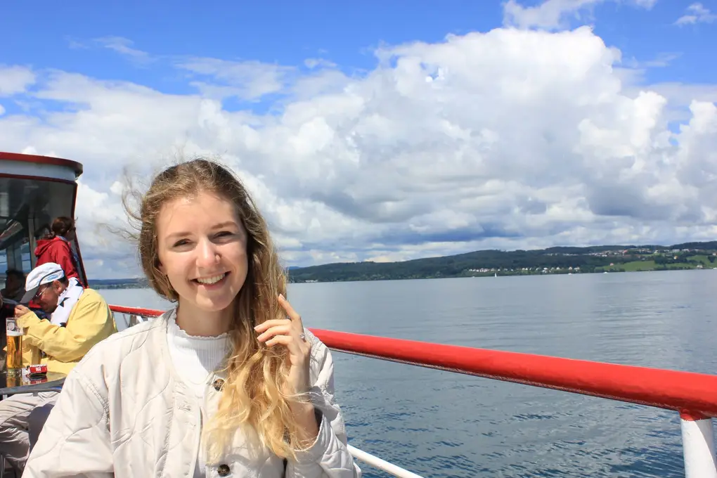 Rebecca lacht an der roten Reling eines Ausflugsschiffs, der Wind im Haar, hinter ihr der See, ein bewaldetes Ufer und große Wolken