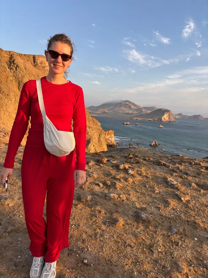Rebecca in Rot vor goldbraunen Steilklippen, dahinter das Meer mit kleinen Felsinseln und eine Halbinsel