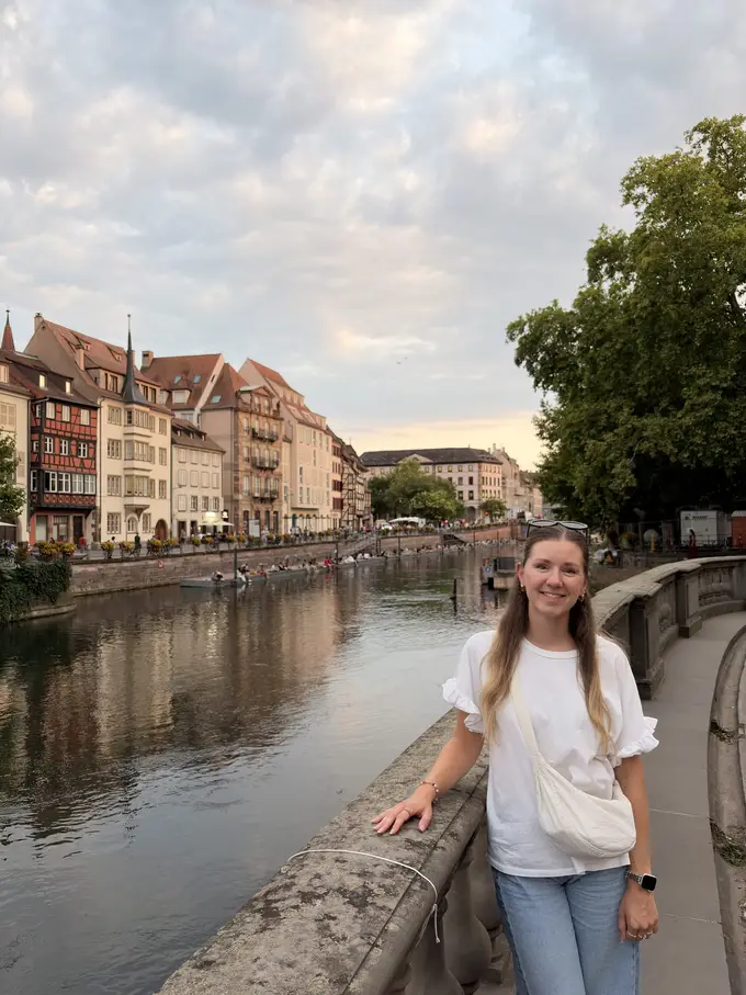 Rebecca lehnt an einer steinernen Brüstung am Fluss, gegenüber eine Häuserzeile mit Fachwerk und Türmchen unter einem Abendhimmel