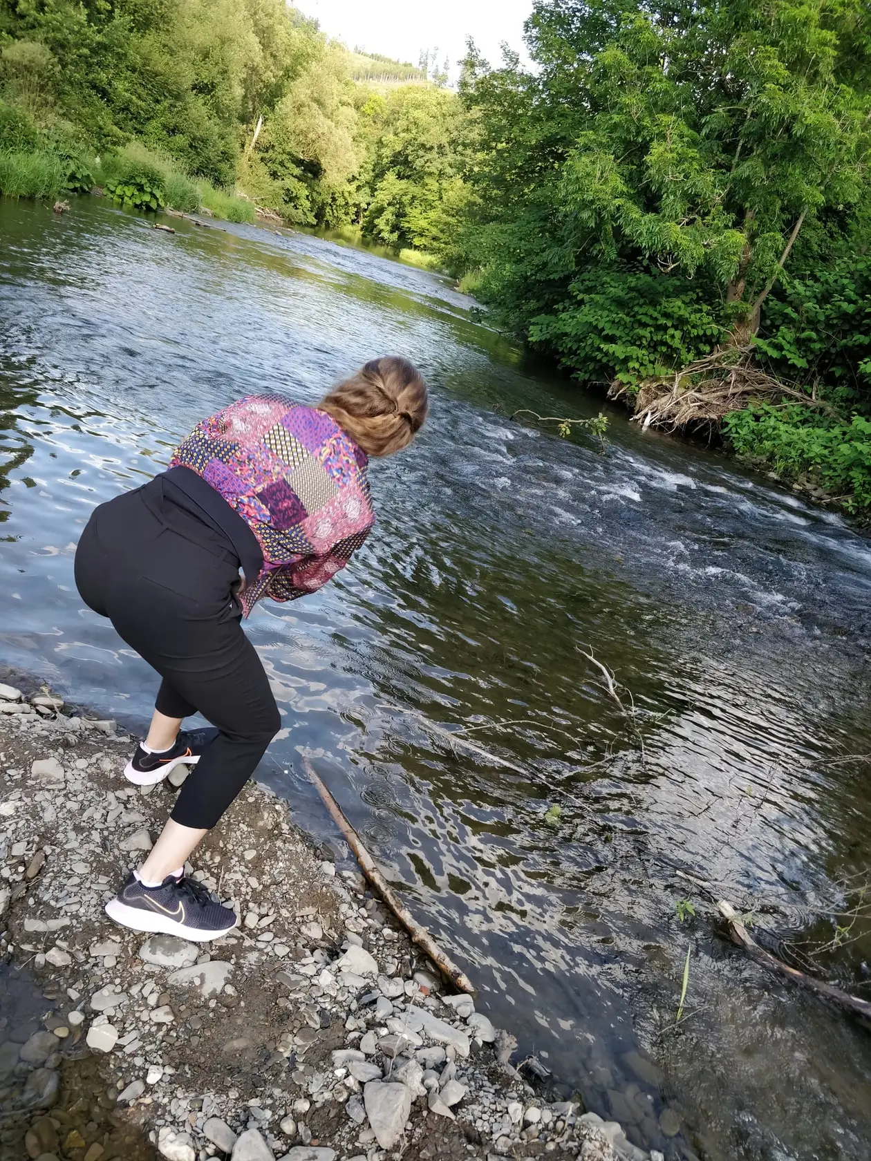 Rebecca beugt sich am steinigen Ufer der Ruhr zum Wasser hinunter, dahinter fließt der Fluss zwischen dicht bewachsenen Ufern