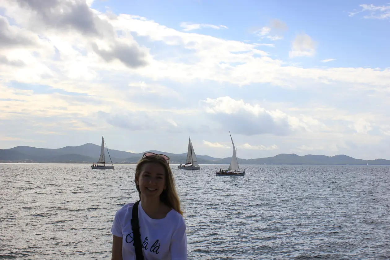 Rebecca im weißen T-Shirt mit Kameragurt am Wasser, hinter ihr das Meer mit Segelbooten und flachen Inseln