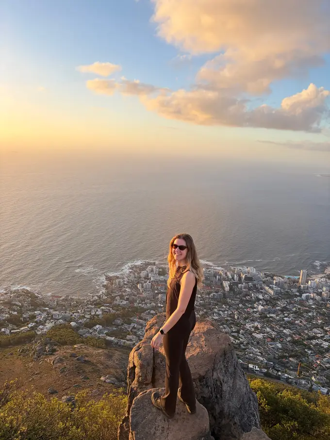 Rebecca steht auf einem Felsen oben auf dem Lion's Head, im Abendlicht auf Meer und Küste blickend