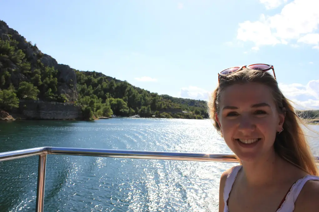 Rebecca an der Reling eines Boots auf dem Fluss, hinter ihr Kielwasser und ein felsiger, bewaldeter Hang