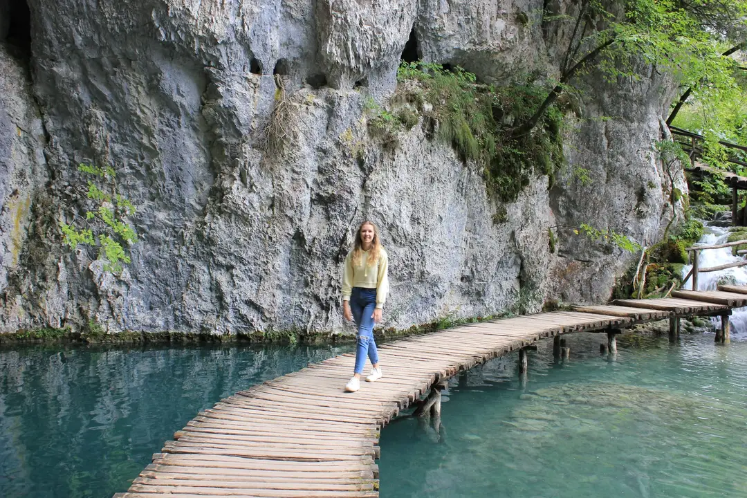Rebecca geht über einen schmalen Holzsteg ohne Geländer, darunter türkises Wasser, dahinter eine hohe graue Felswand