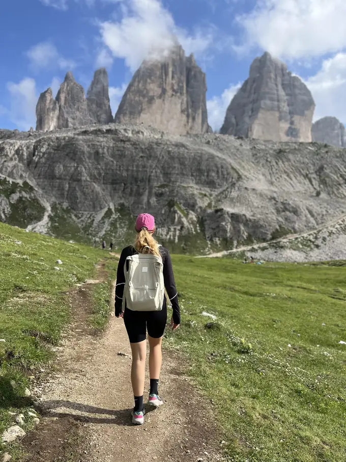 Rebecca von hinten mit pinker Kappe und hellem Rucksack auf einem schmalen Pfad durch eine Almwiese, vor ihr die Drei Zinnen