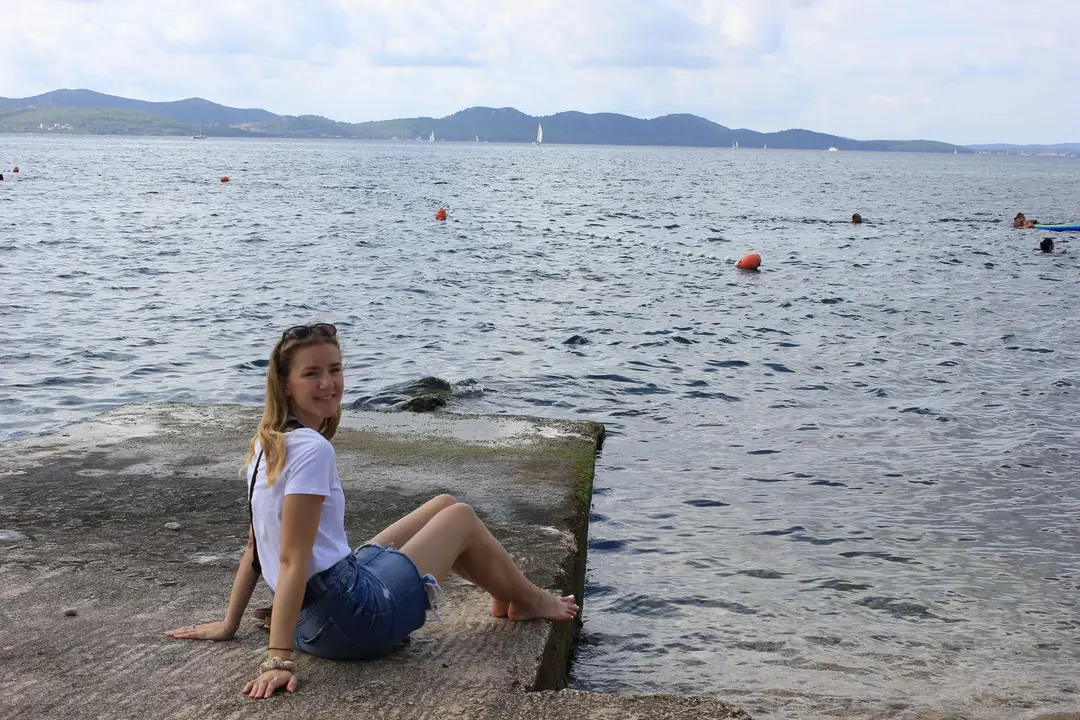 Rebecca sitzt barfuß auf einer Betonplatte am Wasser, hinter ihr eine Bojenleine, Schwimmer und bewaldete Inseln