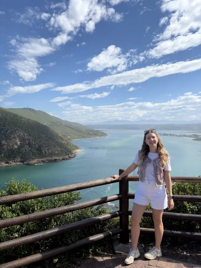 Rebecca steht an einem Holzgeländer mit Blick auf die grüne Landzunge und das Meer bei den Knysna Heads