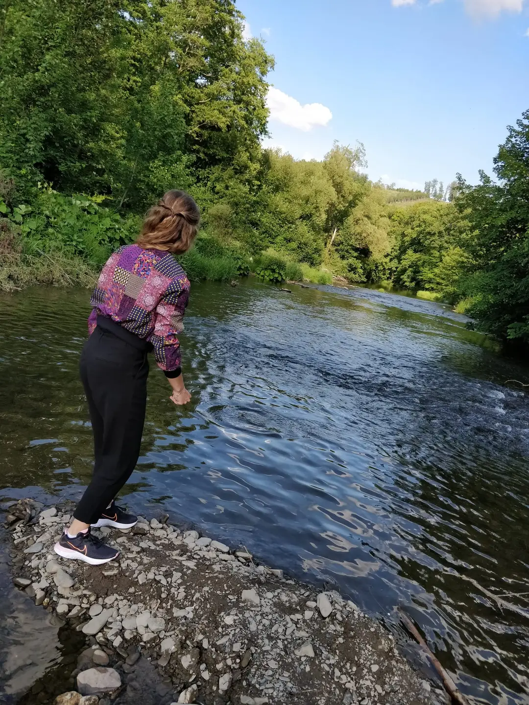 Rebecca steht in bunter Jacke auf einer Kiesbank an der Ruhr und holt mit tief gehaltenem Arm zum Wurf übers Wasser aus, am anderen Ufer dichter Laubwald
