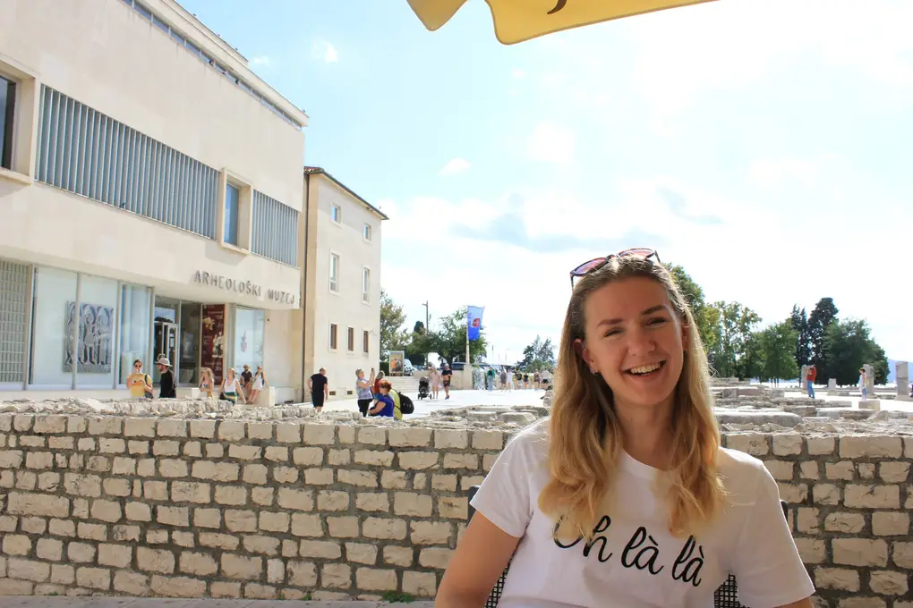 Rebecca lacht an einem Cafétisch, hinter ihr Mauerreste des Forums und ein moderner Bau