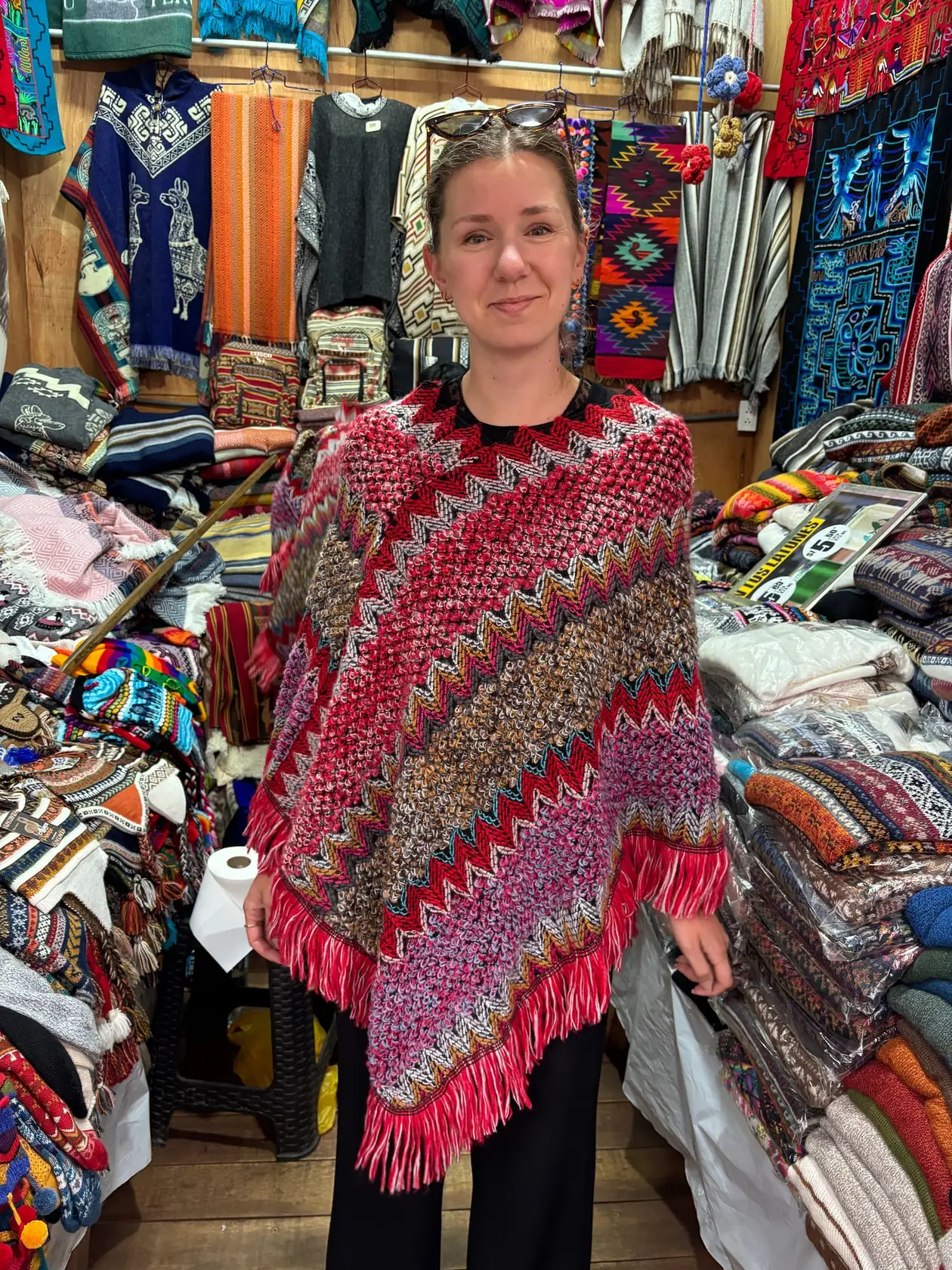 Rebecca probiert an einem Marktstand voller Wolldecken und Pullover einen rot gemusterten Poncho mit Fransen an