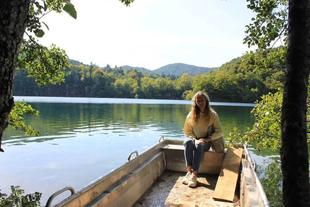 Rebecca sitzt in einem Aluminiumboot am Ufer, dahinter ein ruhiger See und bewaldete Hügel