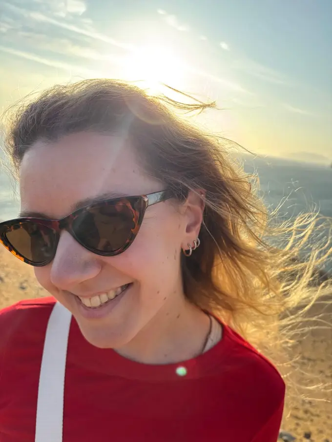 Rebecca mit Sonnenbrille lacht in die Kamera, der Wind weht ihr die Haare ins Gesicht, dahinter die tief stehende Sonne