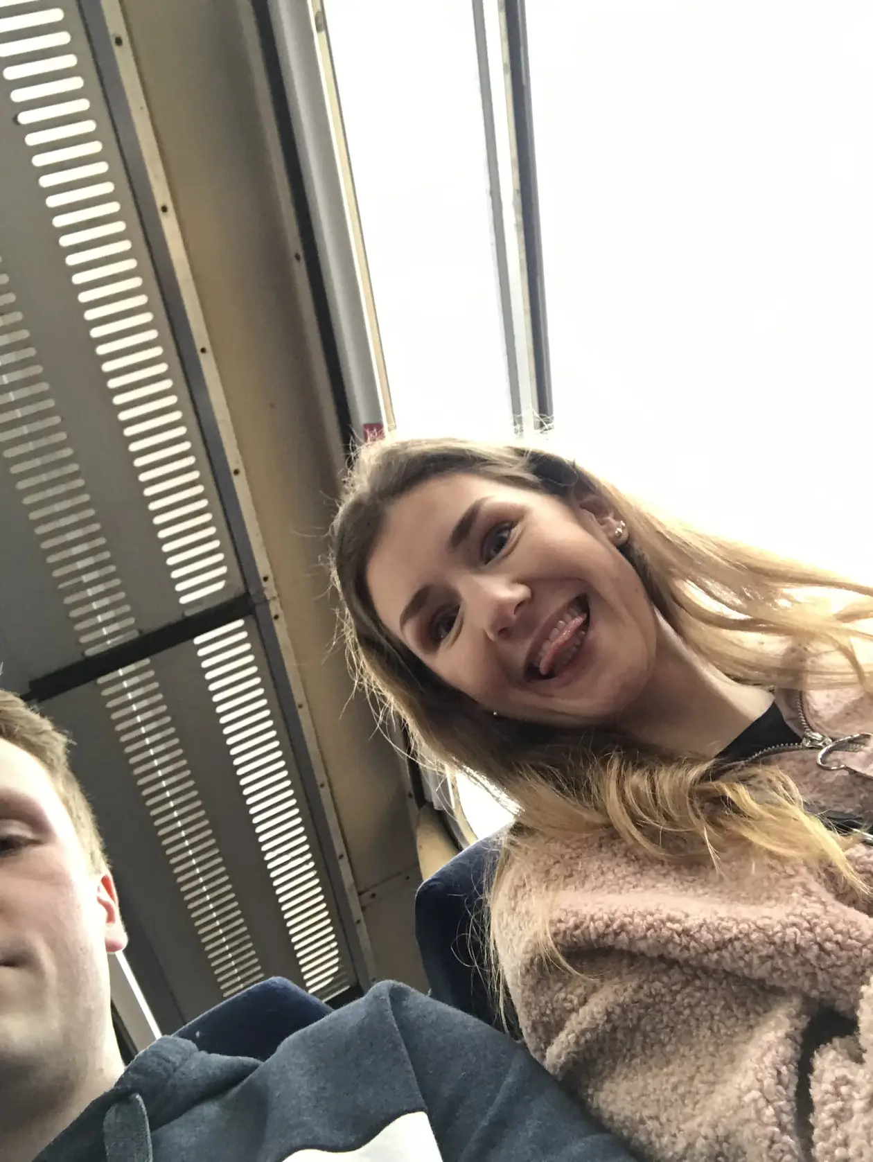 Rebecca sitzt im Zug am Fenster und streckt die Zunge heraus, vom linken Bildrand schaut Max ins Bild