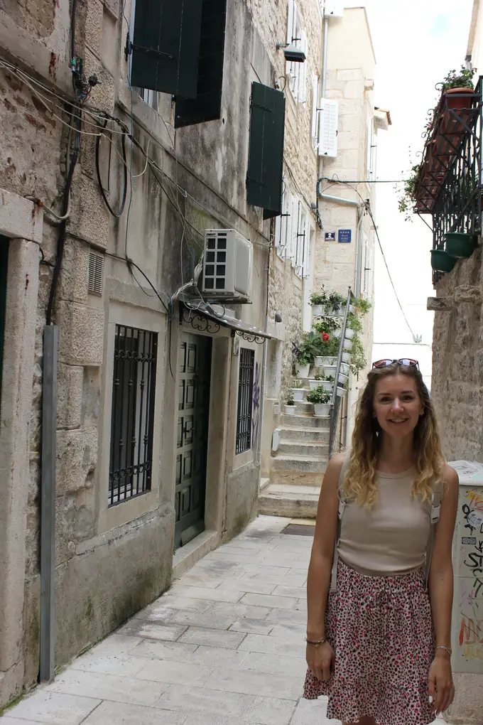 Rebecca in einer engen Gasse zwischen alten Steinhäusern mit Fensterläden, hinter ihr eine Treppe voller Blumentöpfe
