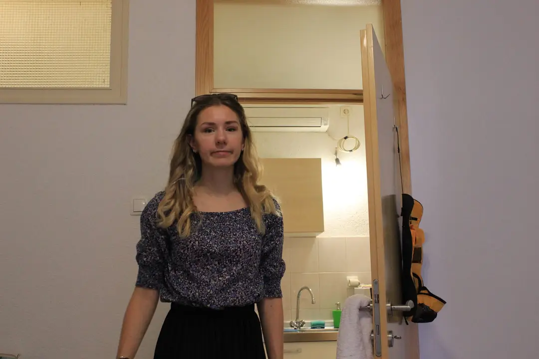 Rebecca steht in einem Zimmer mit hellgrauen Wänden, hinter ihr eine offene Tür zu einer Kochnische mit Spüle und Klimagerät, an der Türklinke ein Handtuch
