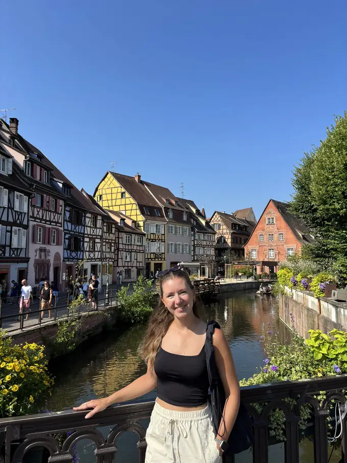 Rebecca lehnt an einem Brückengeländer, hinter ihr ein Kanal und eine Reihe bunter Fachwerkhäuser