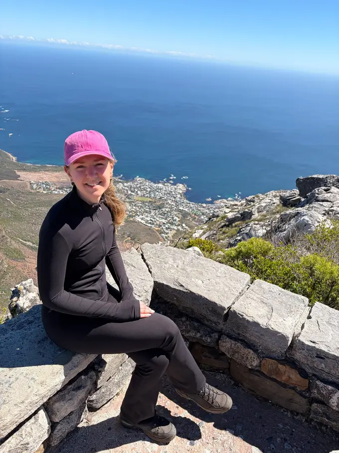 Rebecca sitzt mit pinkfarbener Kappe auf einer Steinmauer oben auf dem Tafelberg, dahinter die Küste