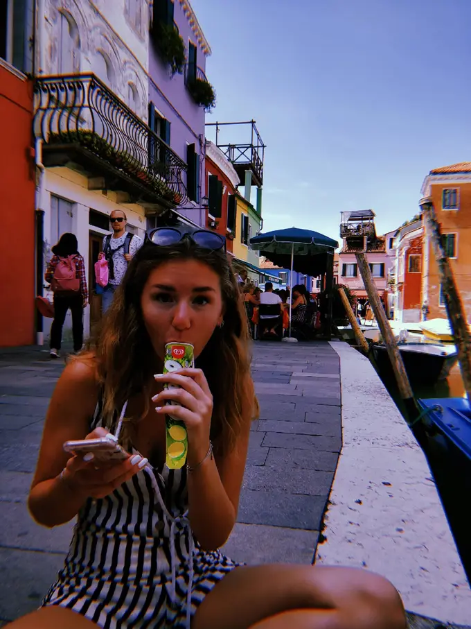 Rebecca sitzt an einer Kaimauer, isst ein Calippo und hält ihr Handy, hinter ihr bunte Häuser und ein Kanal