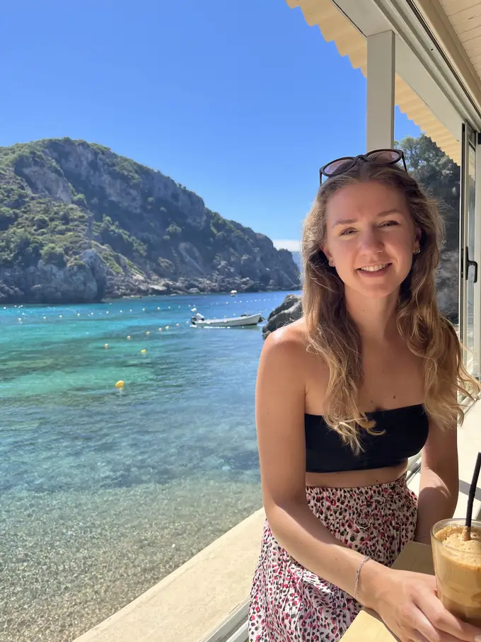 Rebecca an einer Theke mit einem Eiskaffee, hinter ihr klares Wasser und ein bewaldeter Berg